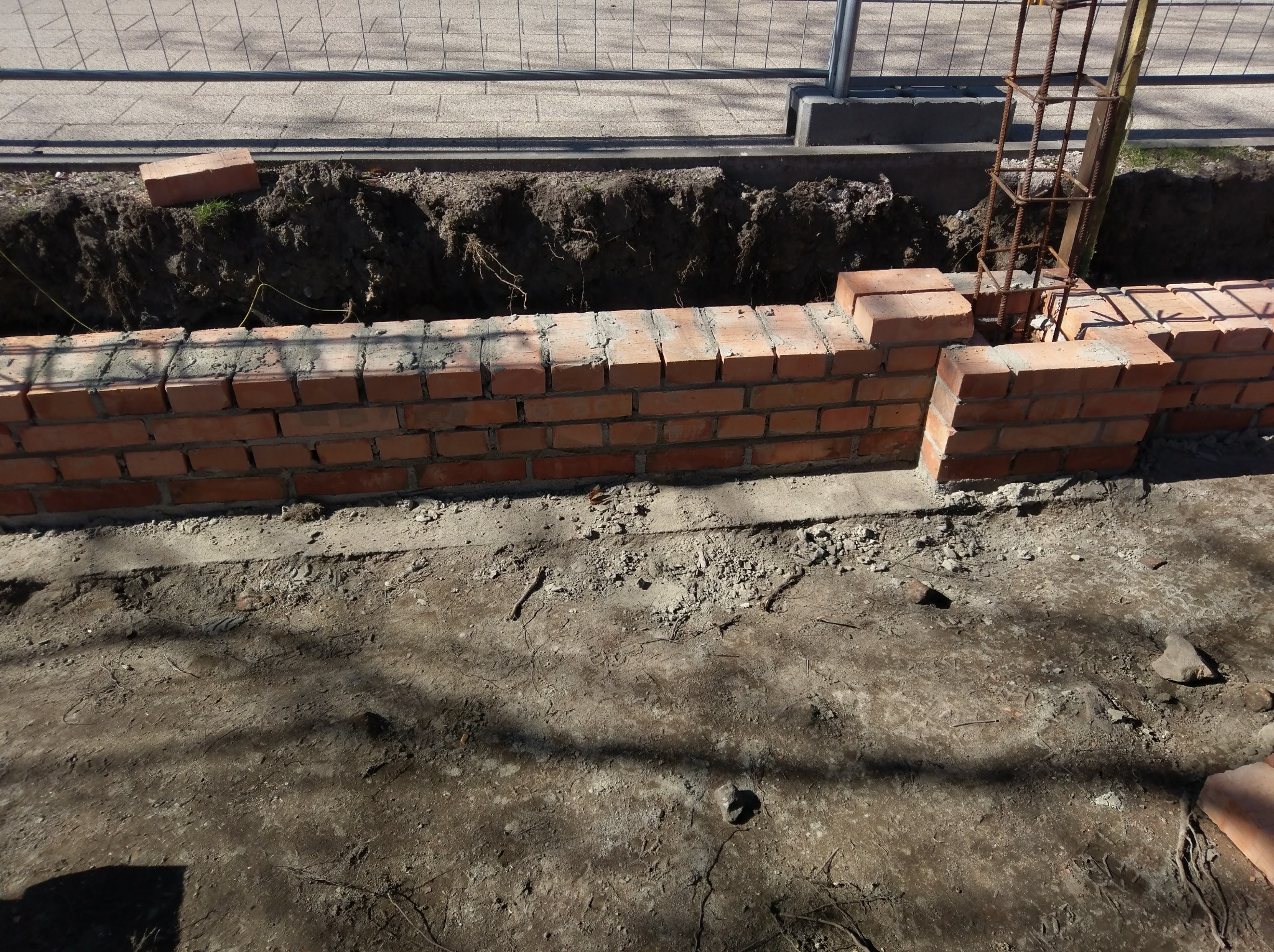 Wznoszenie muru z czerwonej cegły na fundamencie z widocznym zbrojeniem, wykop pod fundament w tle.