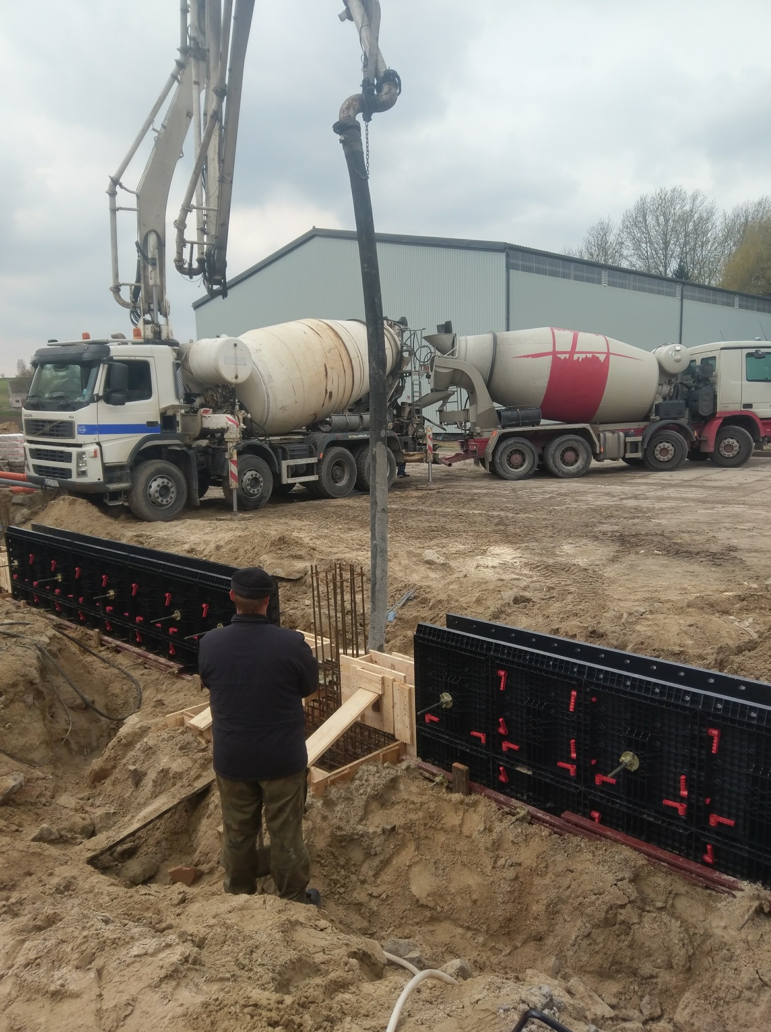 Wylewanie betonu do szalunków fundamentowych za pomocą pompy z betonomieszarki, widoczny pracownik w czapce obserwujący proces.
