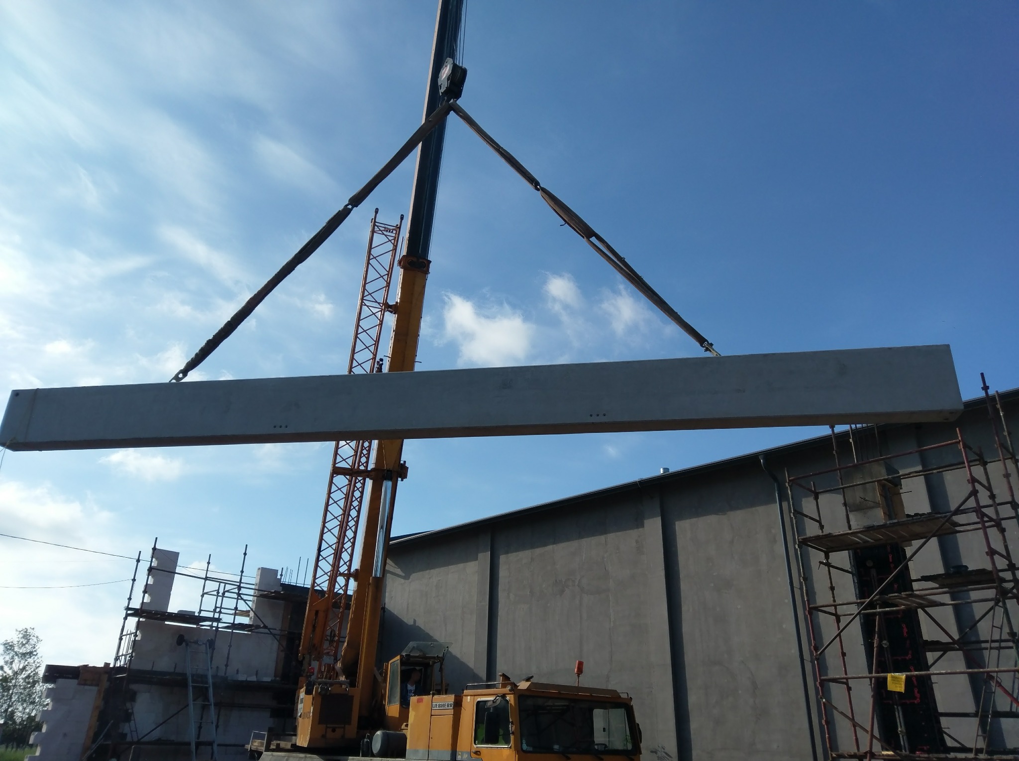 Żółty dźwig Liebherr podnosi betonowy prefabrykat na tle budowanego budynku z szalunkami i rusztowaniami, błękitne niebo z chmurami.