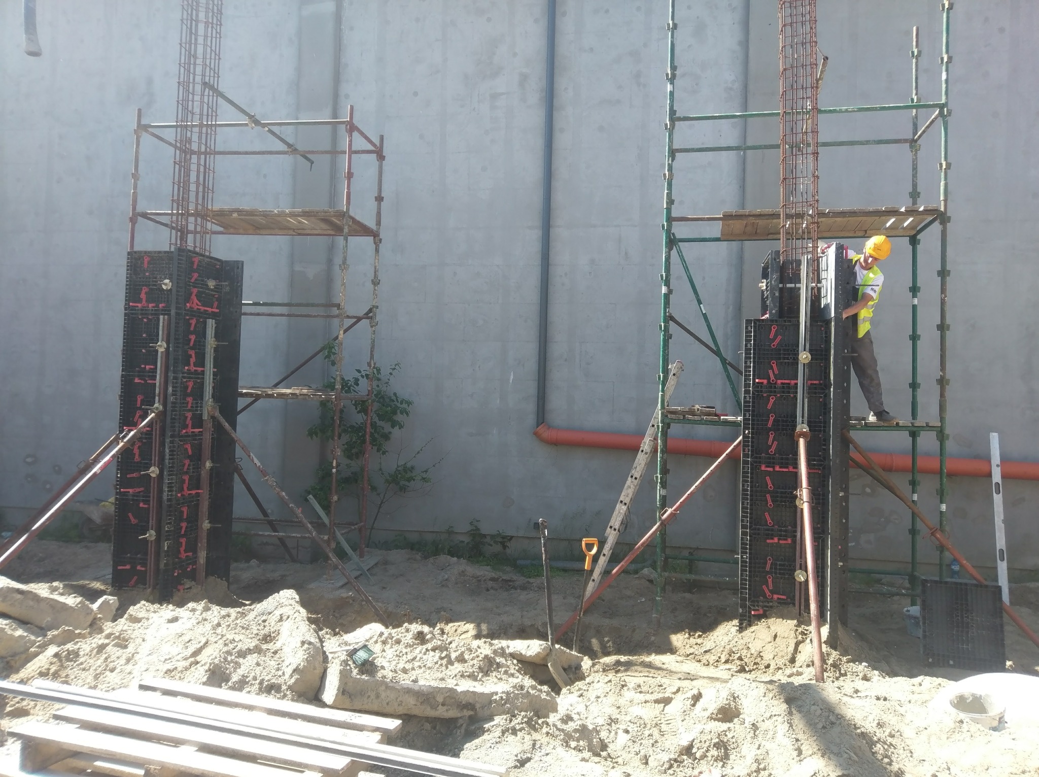 Prace ziemne pod budowę, szalunki kolumnowe zbrojone stalą, ustawione na placu budowy, pracownik w kasku i kamizelce na rusztowaniu, w tle szara ściana budynku.