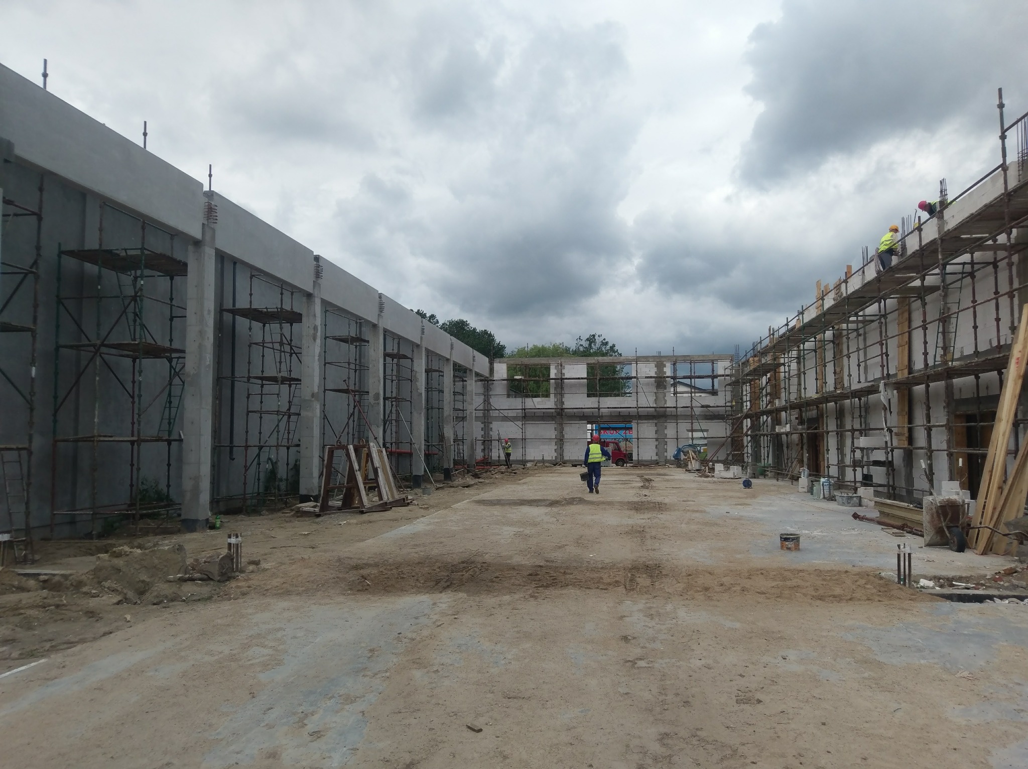 Budowa hali: widok na szkielet budynku z żelbetu, z widocznymi rusztowaniami i pracownikami w kaskach, w tle pochmurne niebo.