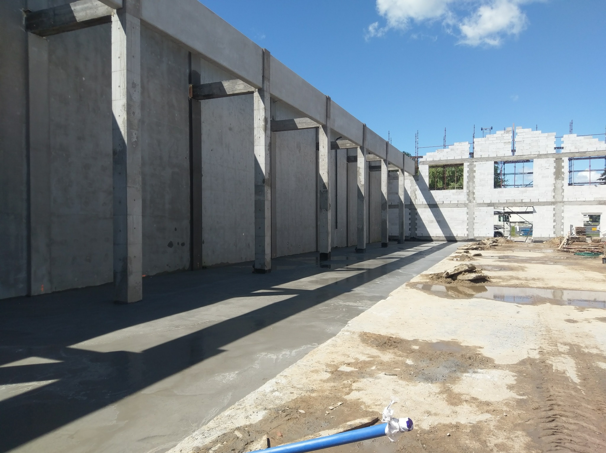 Surowy szkielet budynku żelbetowego w trakcie budowy, widoczne betonowe słupy i belki, świeżo wylana posadzka, częściowo wymurowane ściany z bloczków, błękitne niebo w tle.