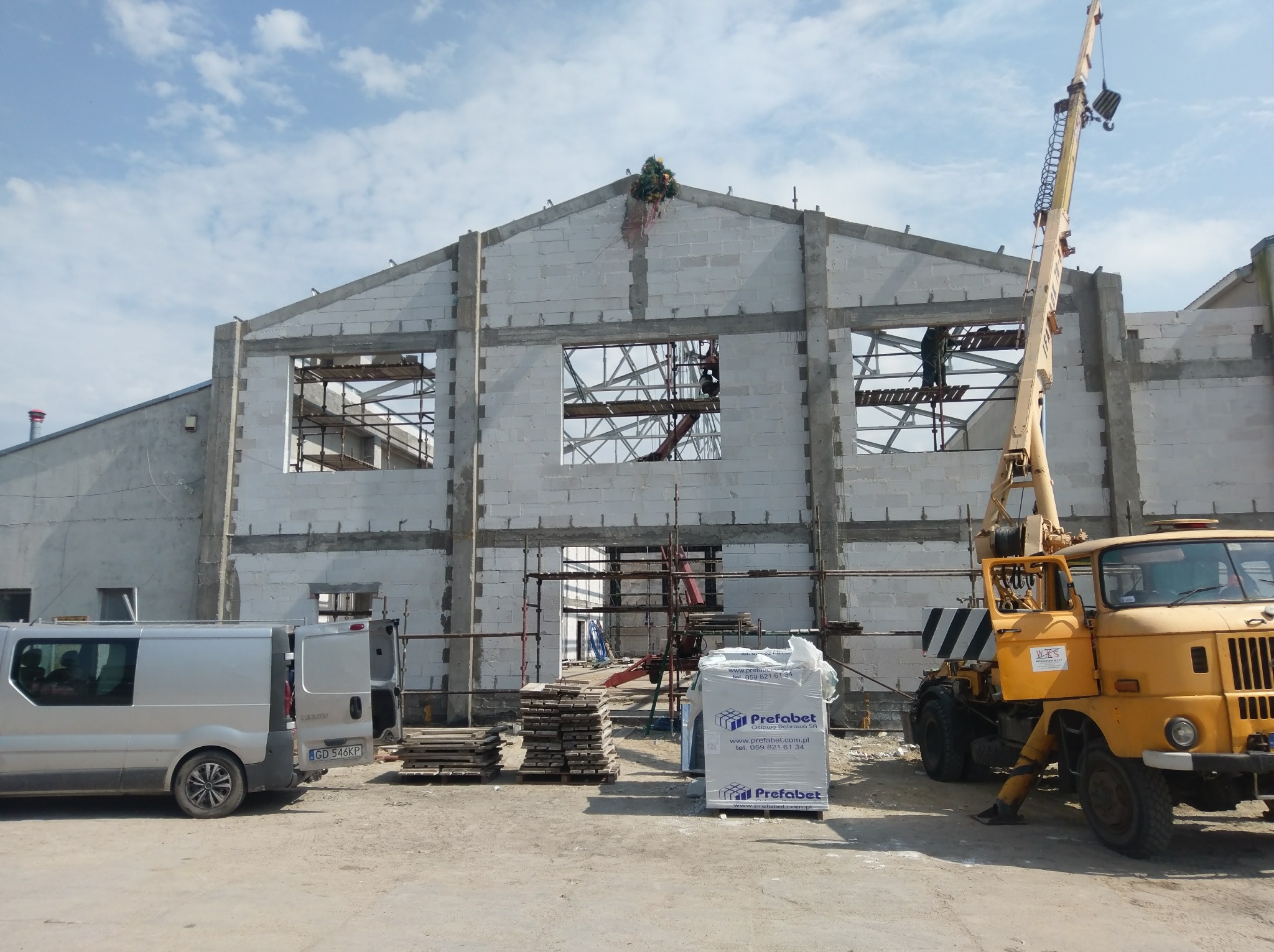 Budowa budynku z białych bloczków, widoczne rusztowania, dźwig, samochód dostawczy i palety z materiałem budowlanym firmy Prefabet, na szczycie budynku wieniec.
