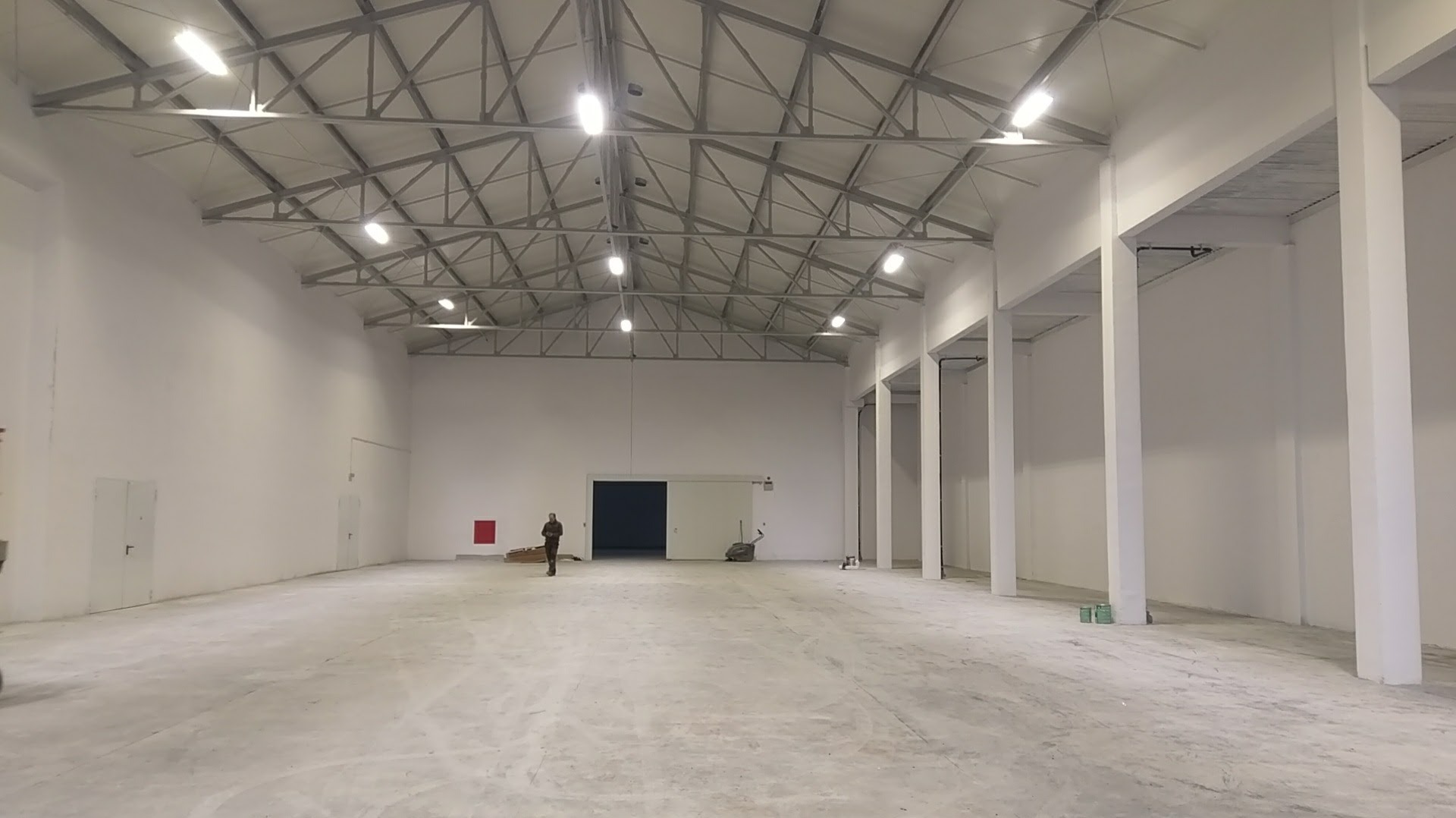 Przestronne wnętrze hali produkcyjnej z żelbetowymi kolumnami, widoczne jasne ściany, betonowa posadzka, stalowa konstrukcja dachu z oświetleniem oraz postać człowieka w oddali przy otwartych wrotach.