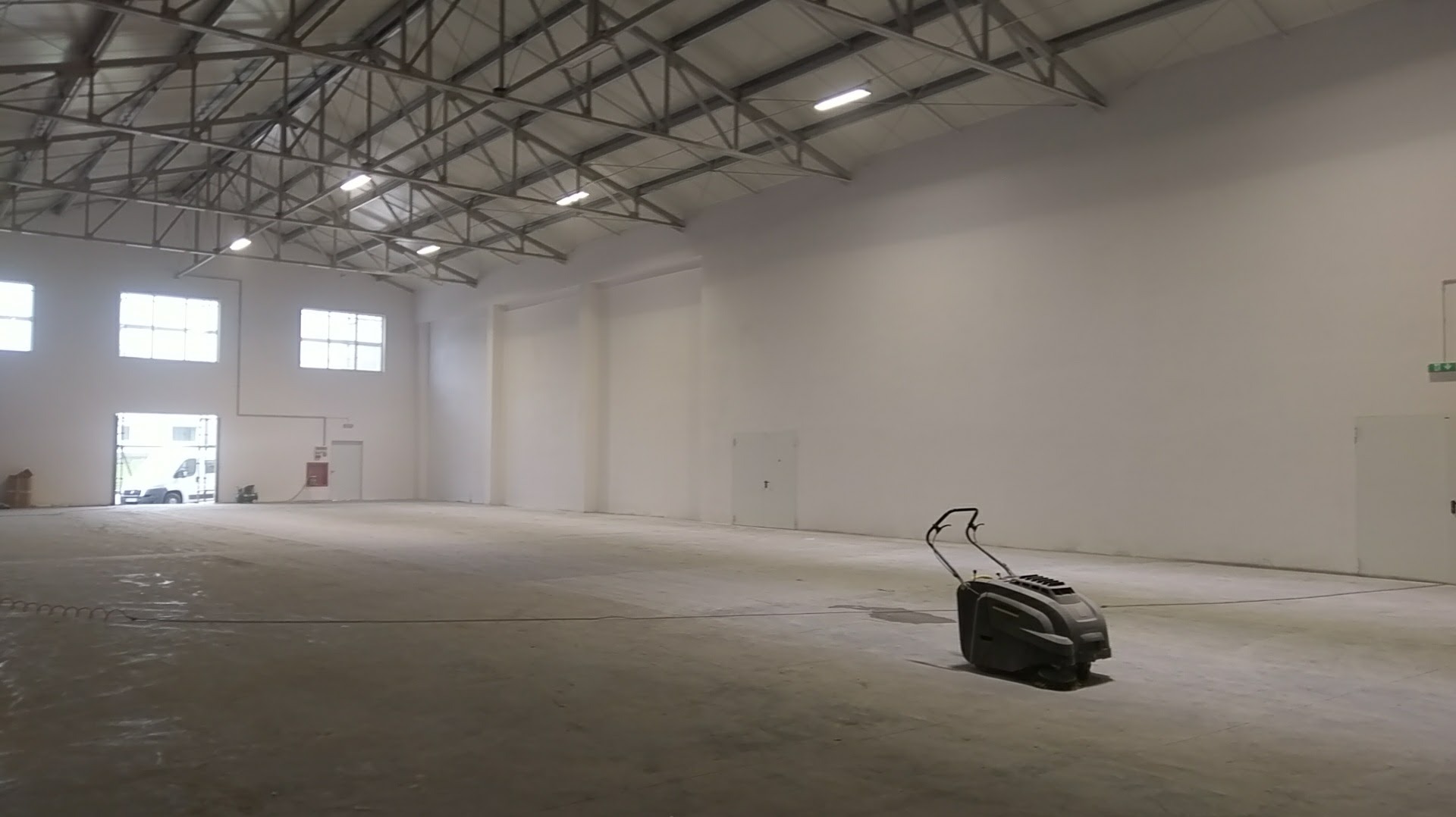 Wnętrze hali magazynowej z betonową posadzką po szlifowaniu, widoczna szlifierka do betonu, ślady po szlifowaniu, okna w tle.