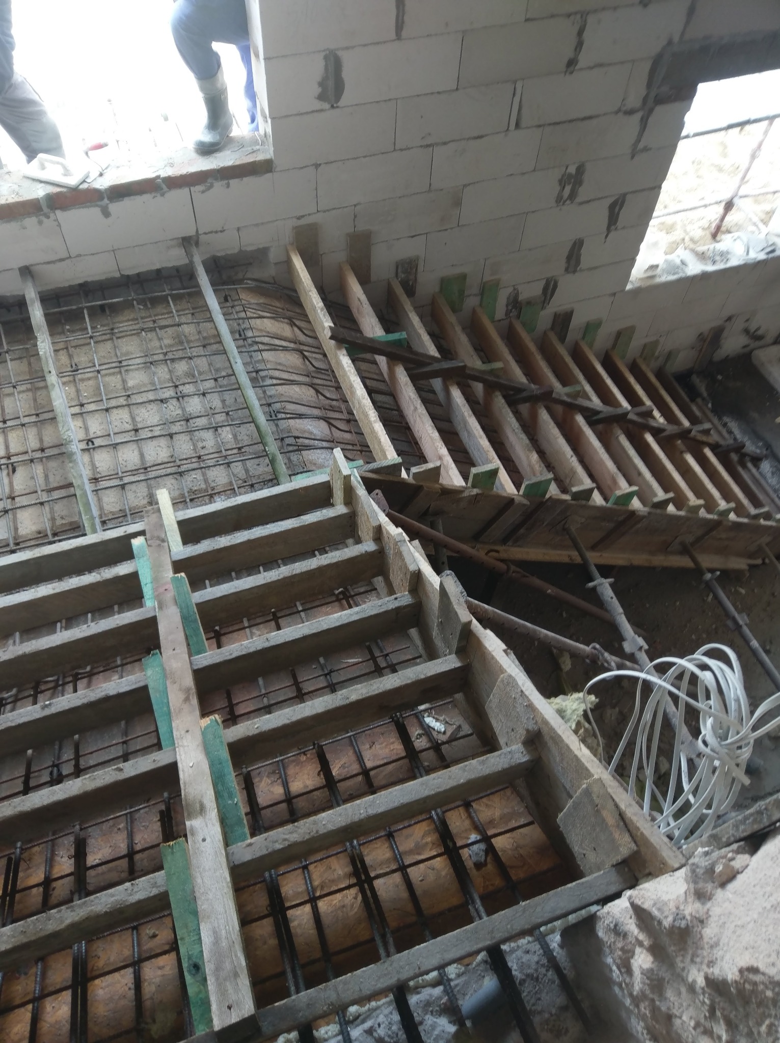 Wykonywanie szalunku i zbrojenia pod fundamenty wewnątrz budynku z białych bloczków, widoczne drewniane stemple i stalowe pręty zbrojeniowe, kabel elektryczny w tle.