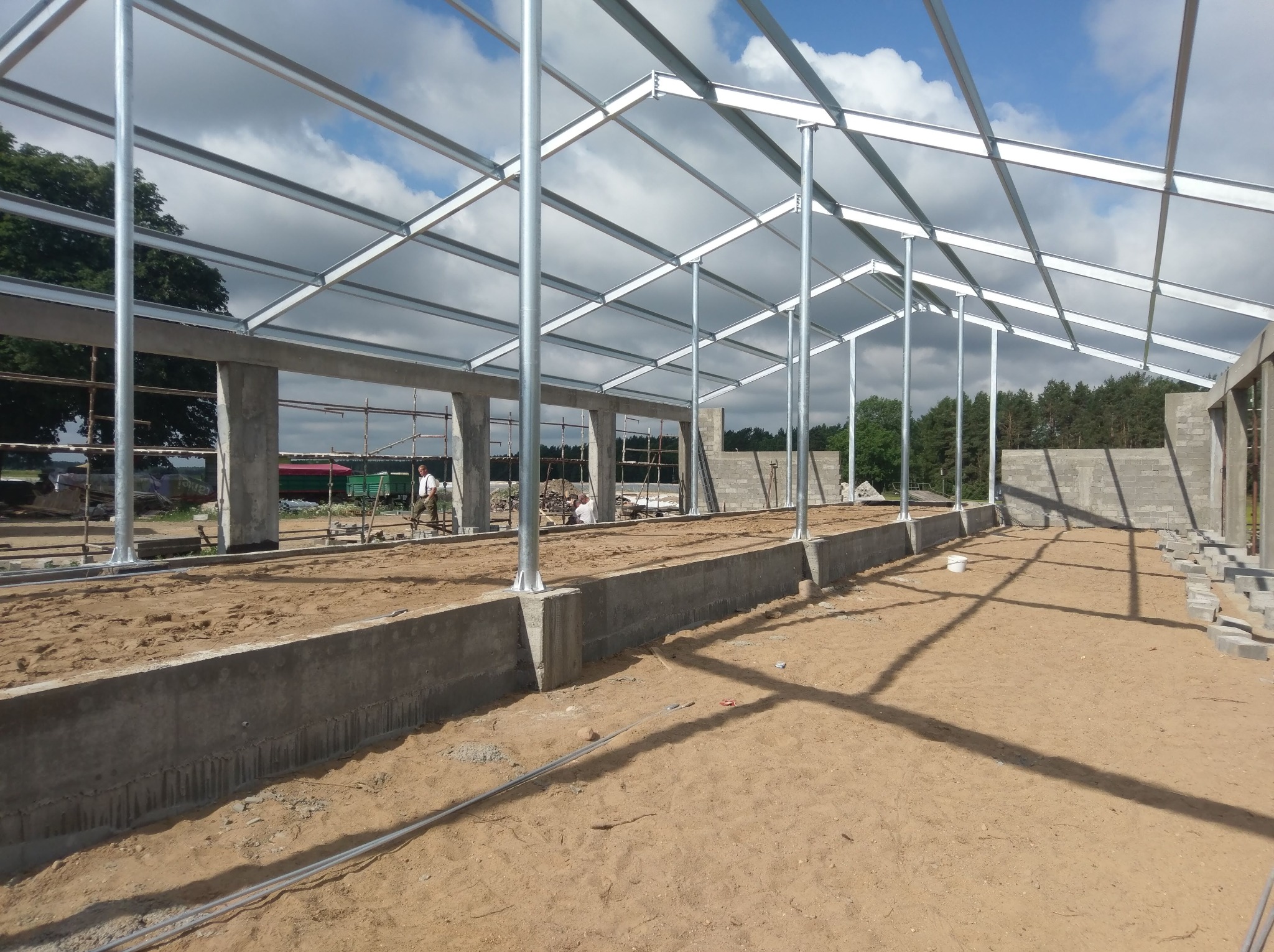 Konstrukcja szkieletowa hali w trakcie budowy, widoczne fundamenty, słupy nośne i stalowa rama dachu, na podłożu piasek, w tle drzewa i błękitne niebo z chmurami.