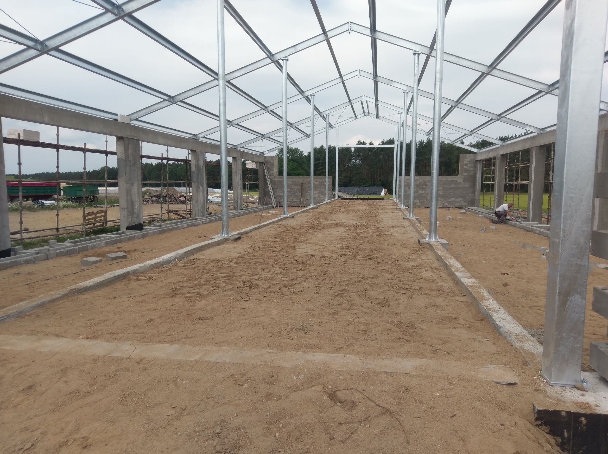 Wnętrze budynku w trakcie budowy z widoczną stalową konstrukcją dachu, surowymi betonowymi ścianami i podłożem z piasku. Robotnik układa bloczki na obrzeżach.