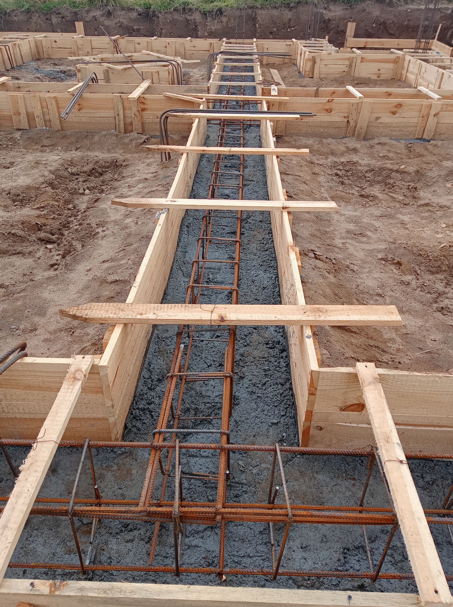 Zalewanie betonem zbrojenia w drewnianym szalunku fundamentu, widok z góry na wykop z ziemią w tle.