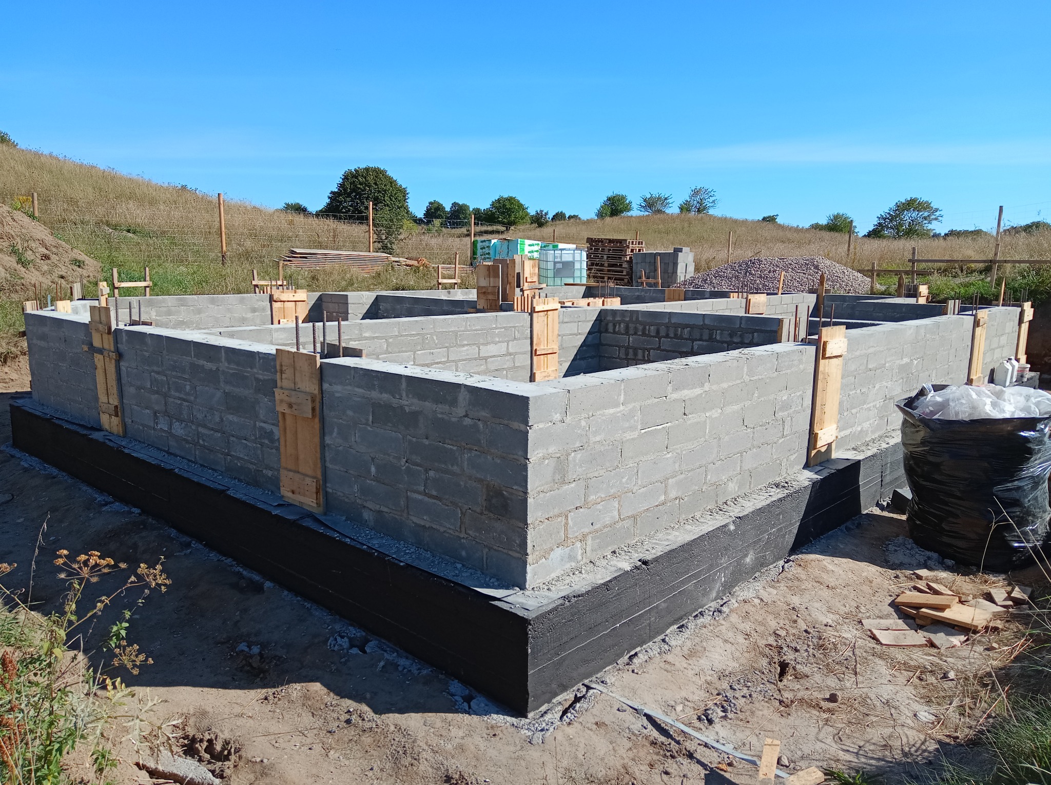 Wzniesione ściany fundamentowe z bloczków betonowych, zabezpieczone czarną izolacją, z drewnianymi stemplami w wykopie na tle łąki i błękitnego nieba.