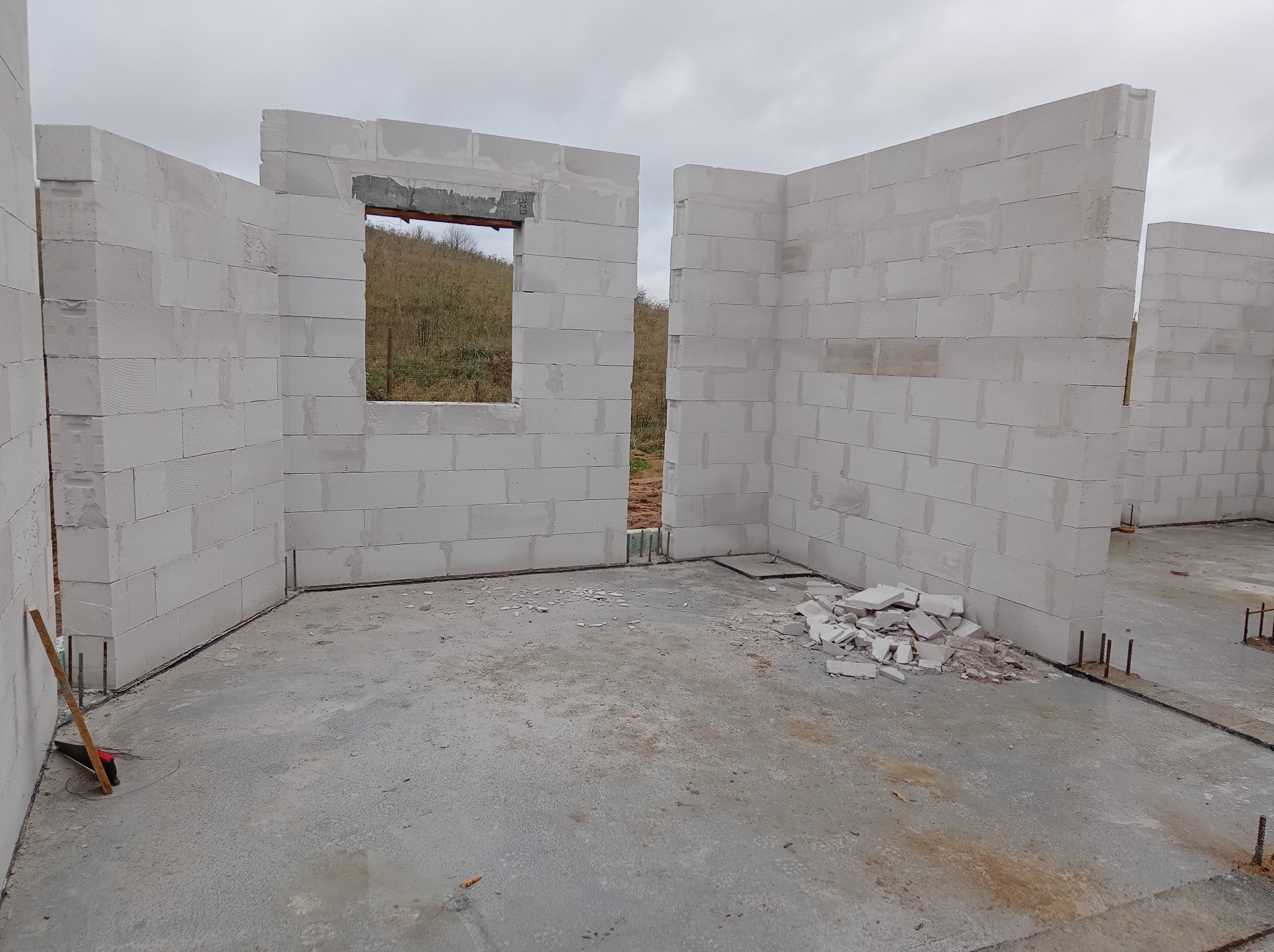 Wnętrze w stanie surowym otwartym z częściowo wzniesionymi ścianami z bloczków, widoczne okno z nadprożem i wystającymi prętami zbrojeniowymi z fundamentu, na posadzce resztki materiałów...