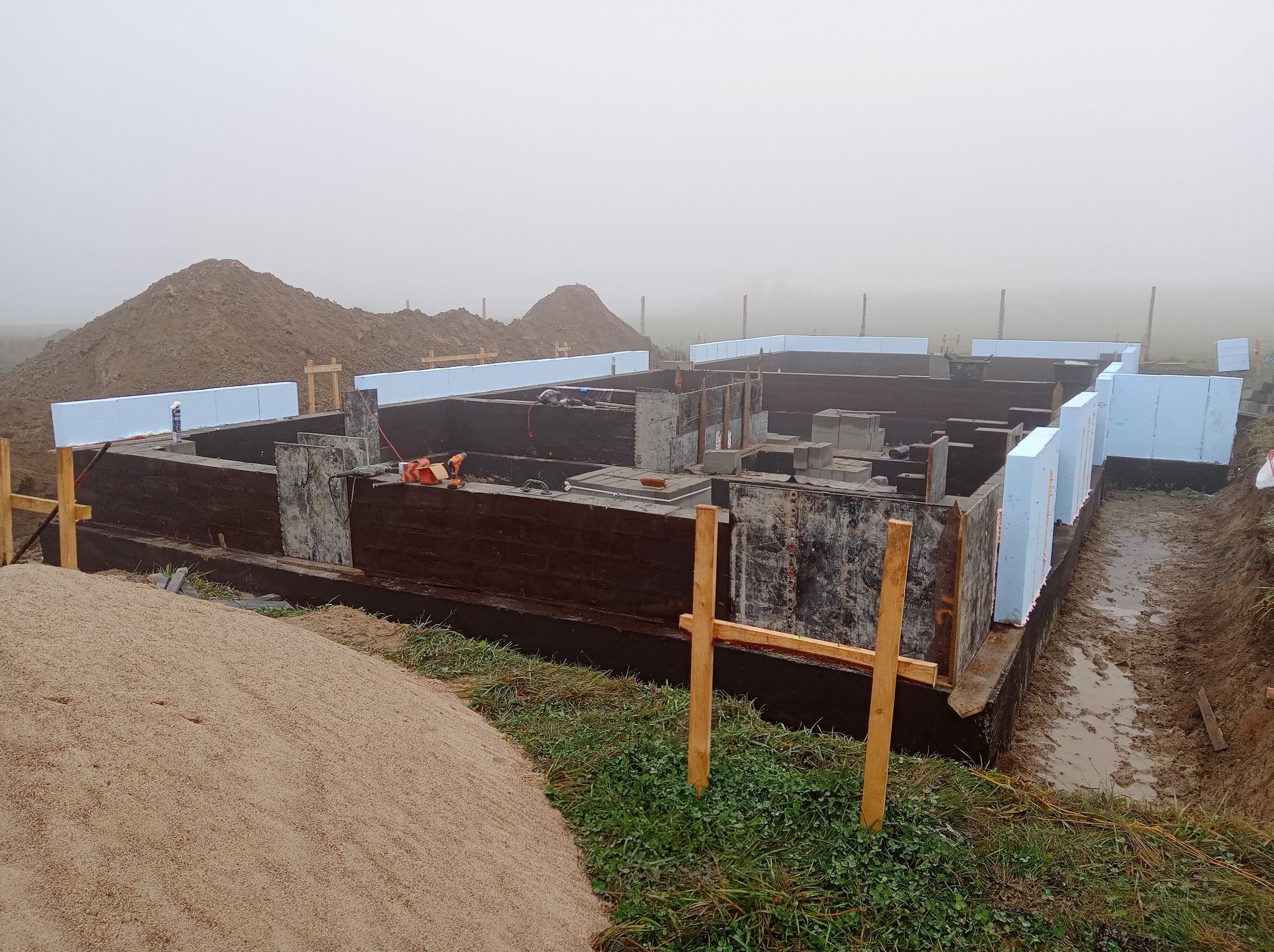 Zalane fundamenty w trakcie budowy, z widocznymi elementami szalunkowymi, izolacją termiczną z polistyrenu ekstrudowanego i stertami ziemi w tle, w pochmurny dzień.