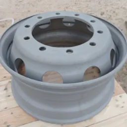 Szara felga samochodowa z otworami na śruby i wentylację, leżąca na drewnianych deskach, z widocznym piaskowym podłożem w tle.