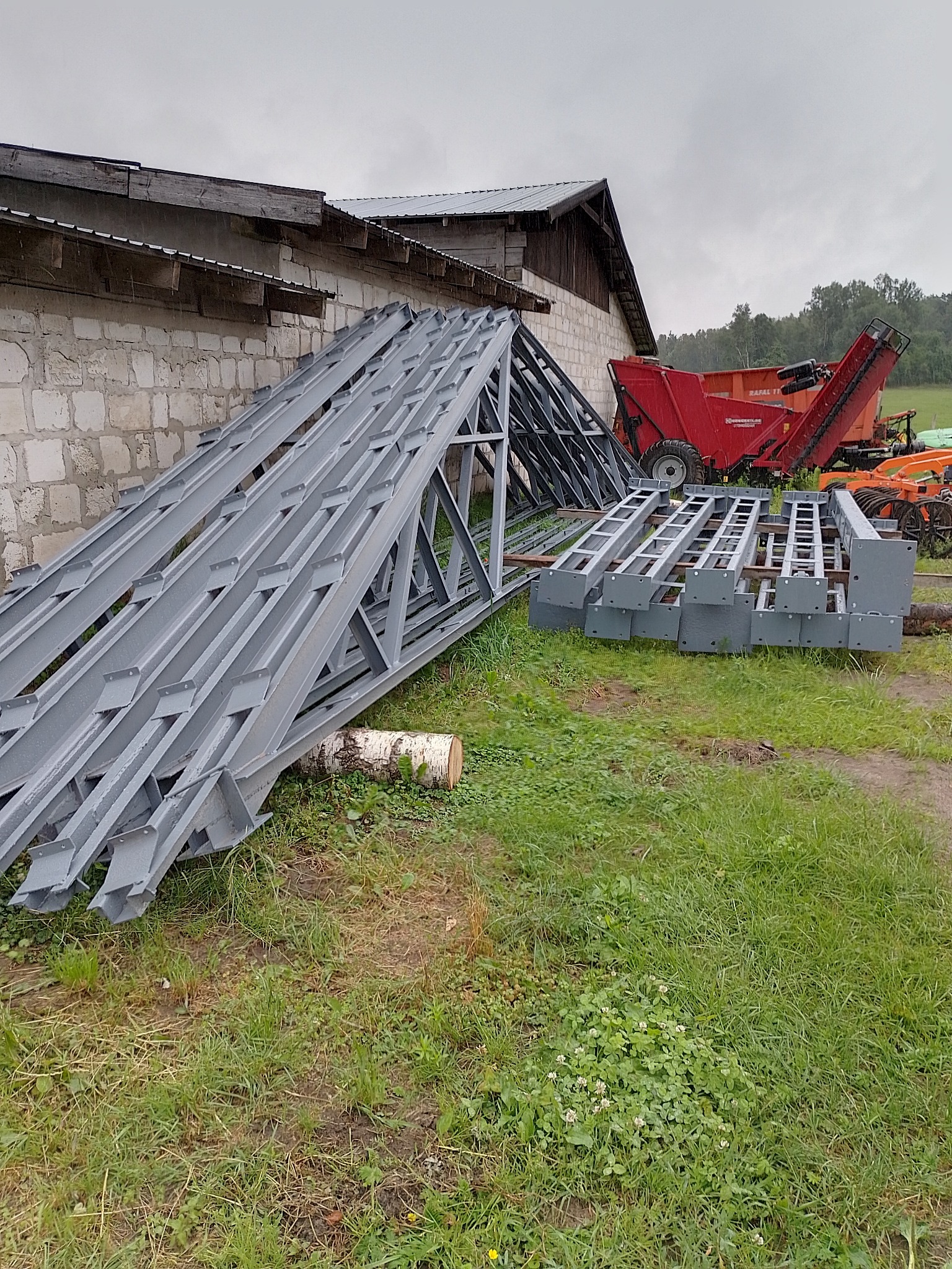 Stalowe elementy konstrukcyjne pomalowane na szaro, leżące na trawie przed budynkiem gospodarczym i czerwonymi maszynami rolniczymi w tle.