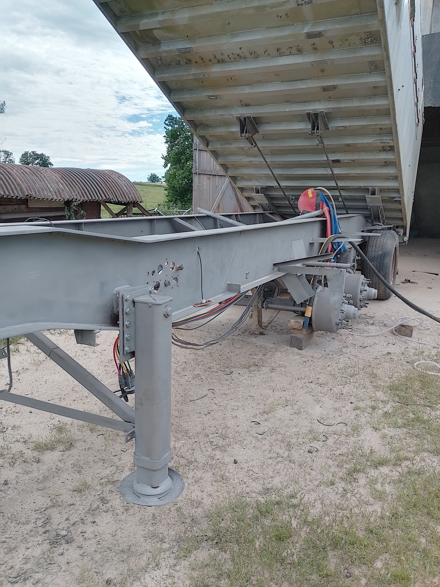 Szara rama naczepy po renowacji, z podniesioną skrzynią ładunkową, widoczne ślady świeżego malowania i elementy hydrauliczne, tło wiejskie z budynkiem gospodarczym.