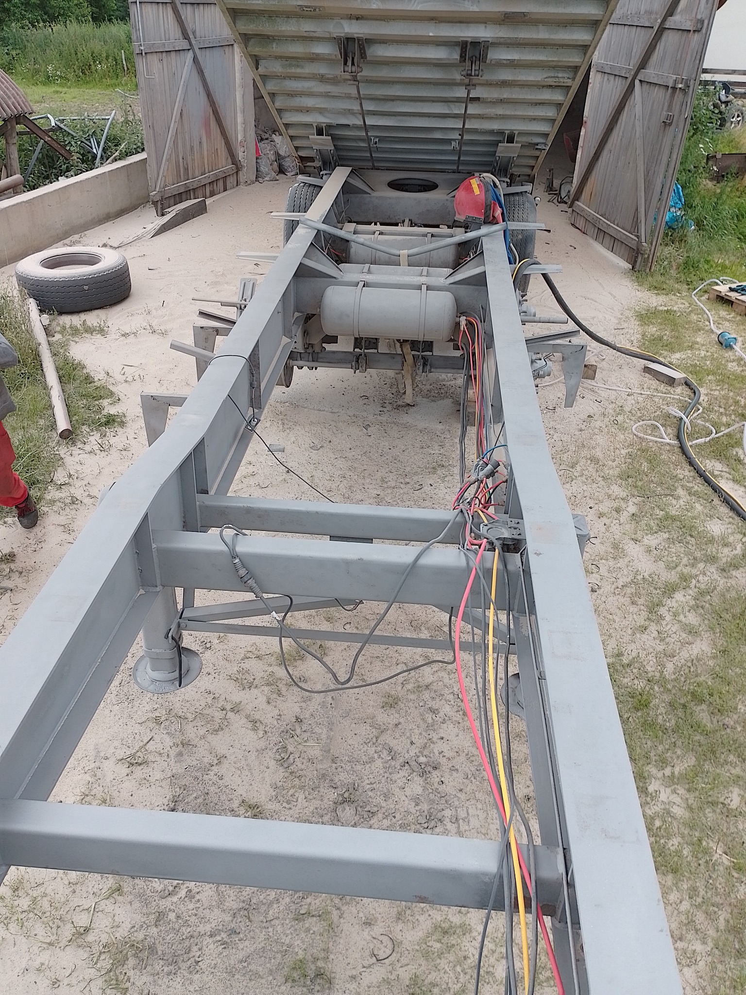 Rama przyczepy po odświeżeniu, prawdopodobnie pomalowana na szaro, widoczne przewody elektryczne, opona i otwarte drzwi skrzyni ładunkowej w tle.