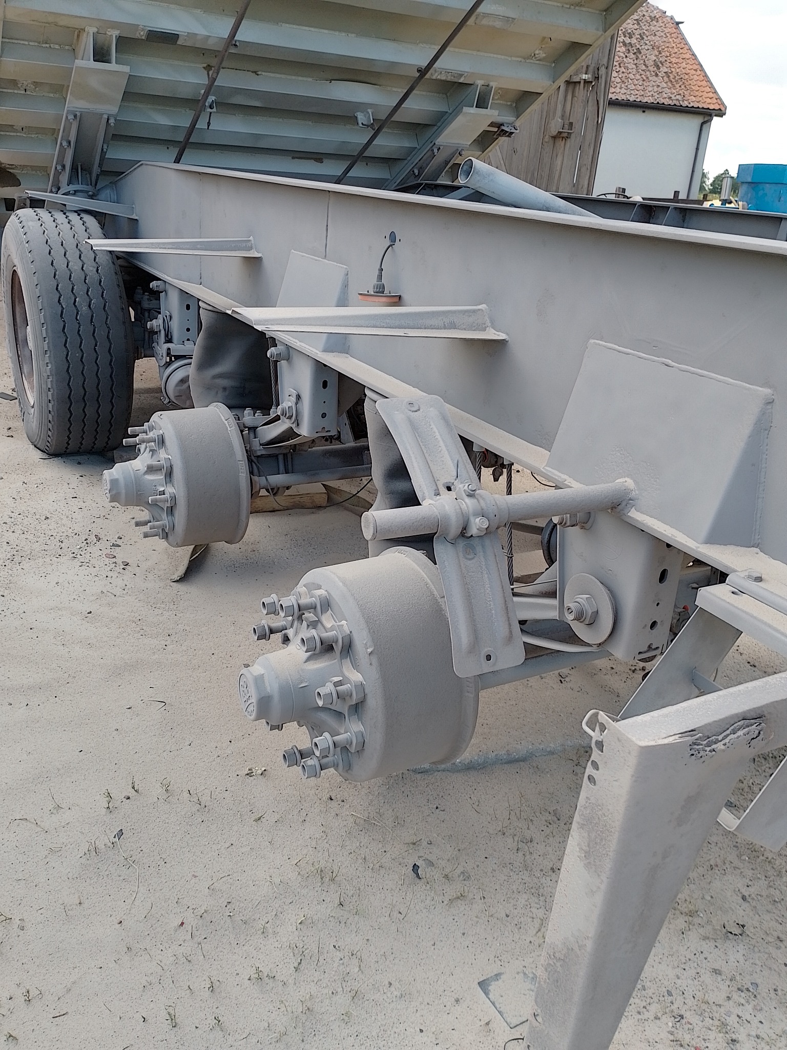 Szare podwozie naczepy po piaskowaniu i malowaniu proszkowym, widoczne elementy zawieszenia, osie i koła, zarys budynku w tle.