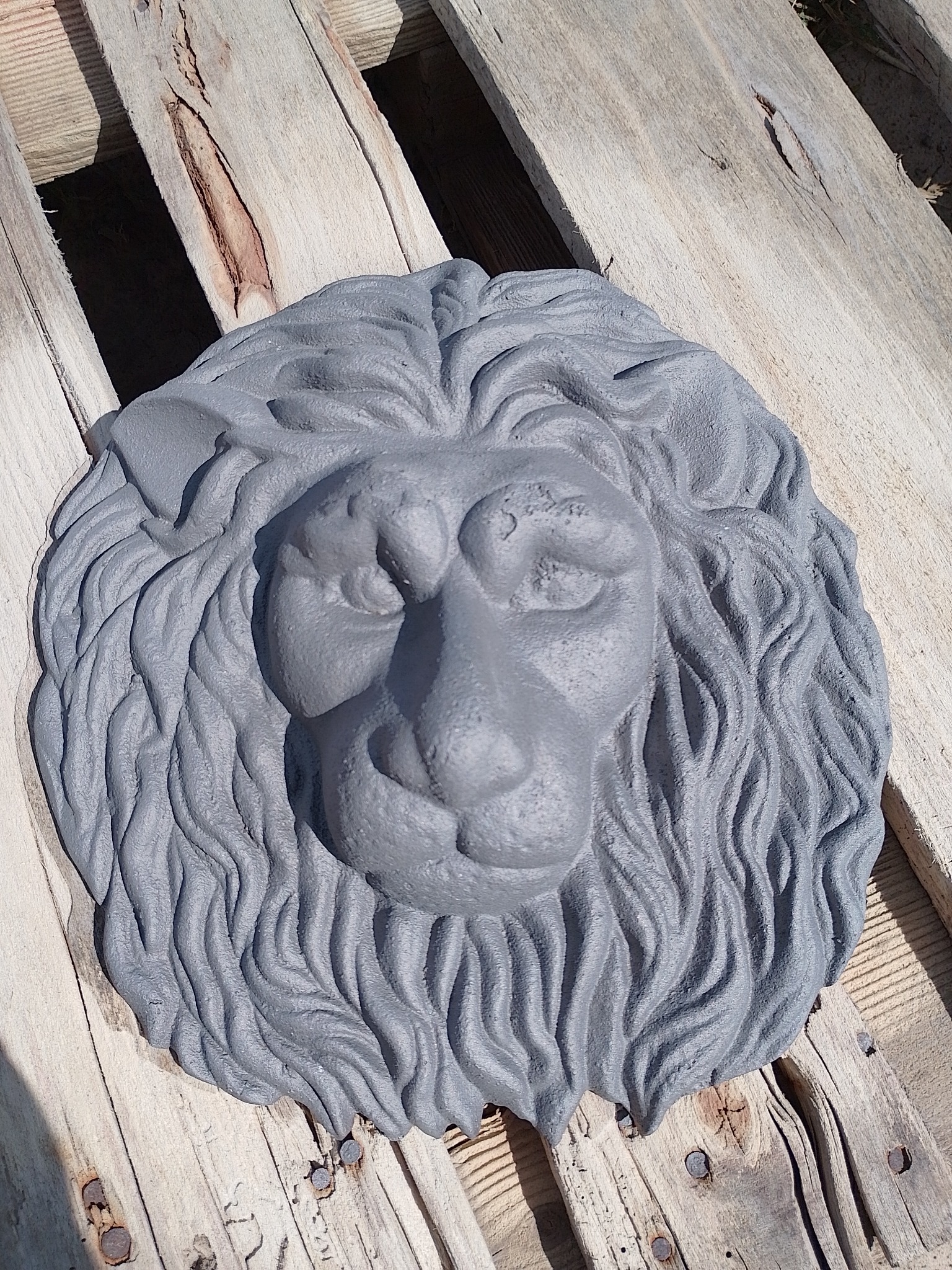 Szara, betonowa głowa lwa z wyraźną fakturą, leżąca na drewnianej palecie w jasnym świetle słonecznym.