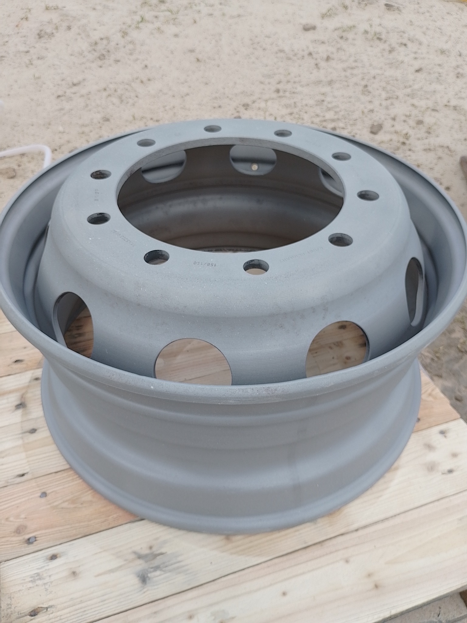 Szara felga samochodowa z otworami na śruby i wentylację, leżąca na drewnianych deskach, z widocznym piaskowym podłożem w tle.