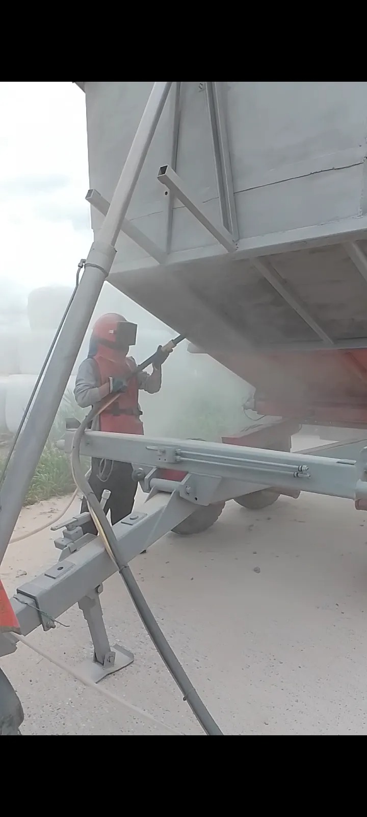 Pracownik w kombinezonie ochronnym oczyszcza strumieniem ścierniwa przyczepę rolniczą, przygotowując ją do malowania.