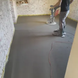 Pracownik w kaloszach i nakładkach na buty wyrównuje świeżo wylaną, szarą posadzkę jastrychową z pomocą zacieraczki mechanicznej w pomieszczeniu z surowymi, ceglanymi ścianami.