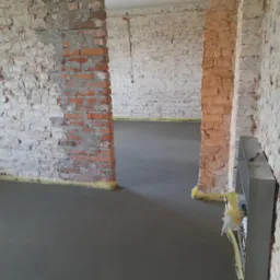 Świeżo wylana, gładka, szara posadzka w remontowanym pomieszczeniu z surowymi, częściowo otynkowanymi ścianami z cegły, zabezpieczonymi żółtą taśmą. Widoczne instalacje elektryczne w ścianie.