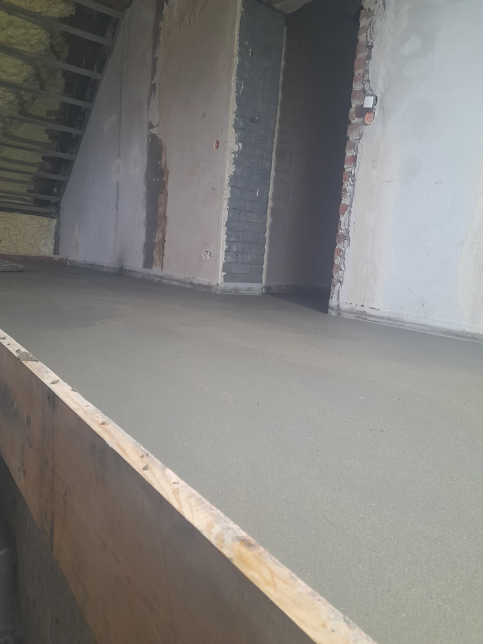 Świeżo wylana, gładka powierzchnia podłogi w trakcie prac wykończeniowych, widoczne surowe ściany i fragment drewnianego szalunku.