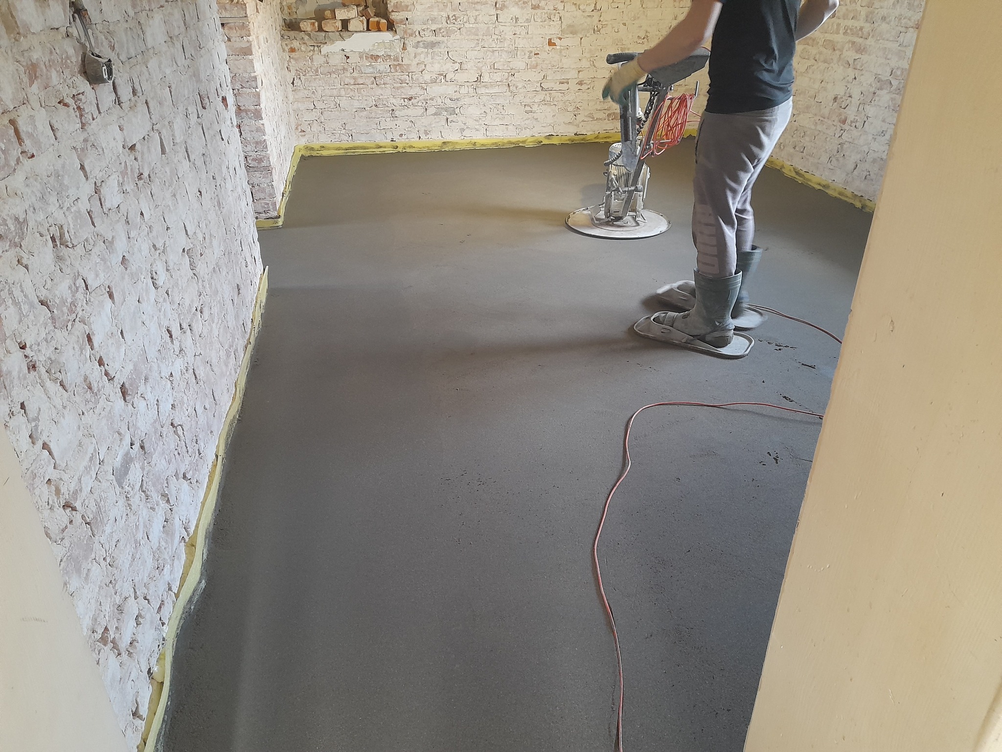 Pracownik w kaloszach i nakładkach na buty wyrównuje świeżo wylaną, szarą posadzkę jastrychową z pomocą zacieraczki mechanicznej w pomieszczeniu z surowymi, ceglanymi ścianami.