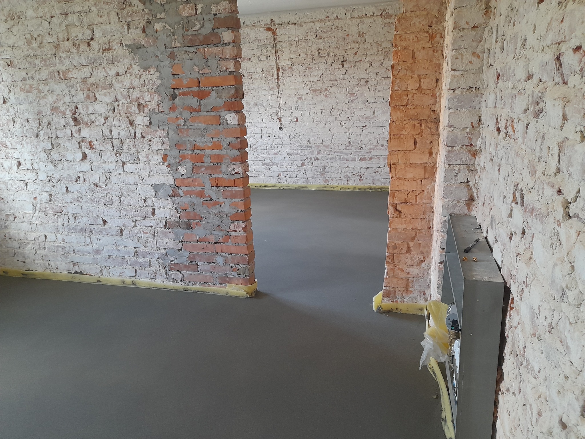 Świeżo wylana, gładka, szara posadzka w remontowanym pomieszczeniu z surowymi, częściowo otynkowanymi ścianami z cegły, zabezpieczonymi żółtą taśmą. Widoczne instalacje elektryczne w ścianie.