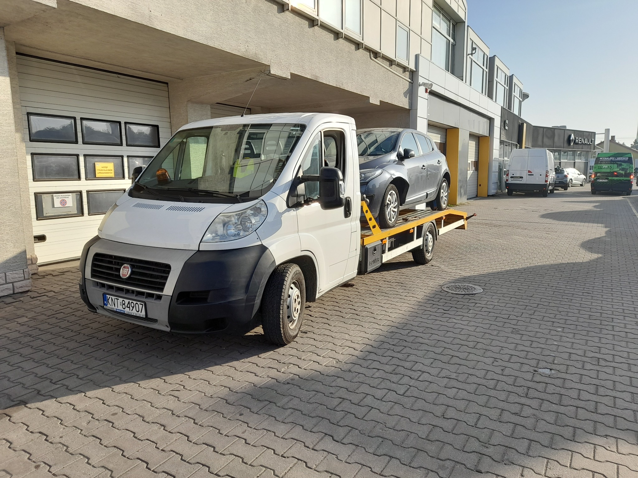 Laweta z szarym samochodem osobowym na tle budynku Renault, sfotografowana na kostce brukowej.