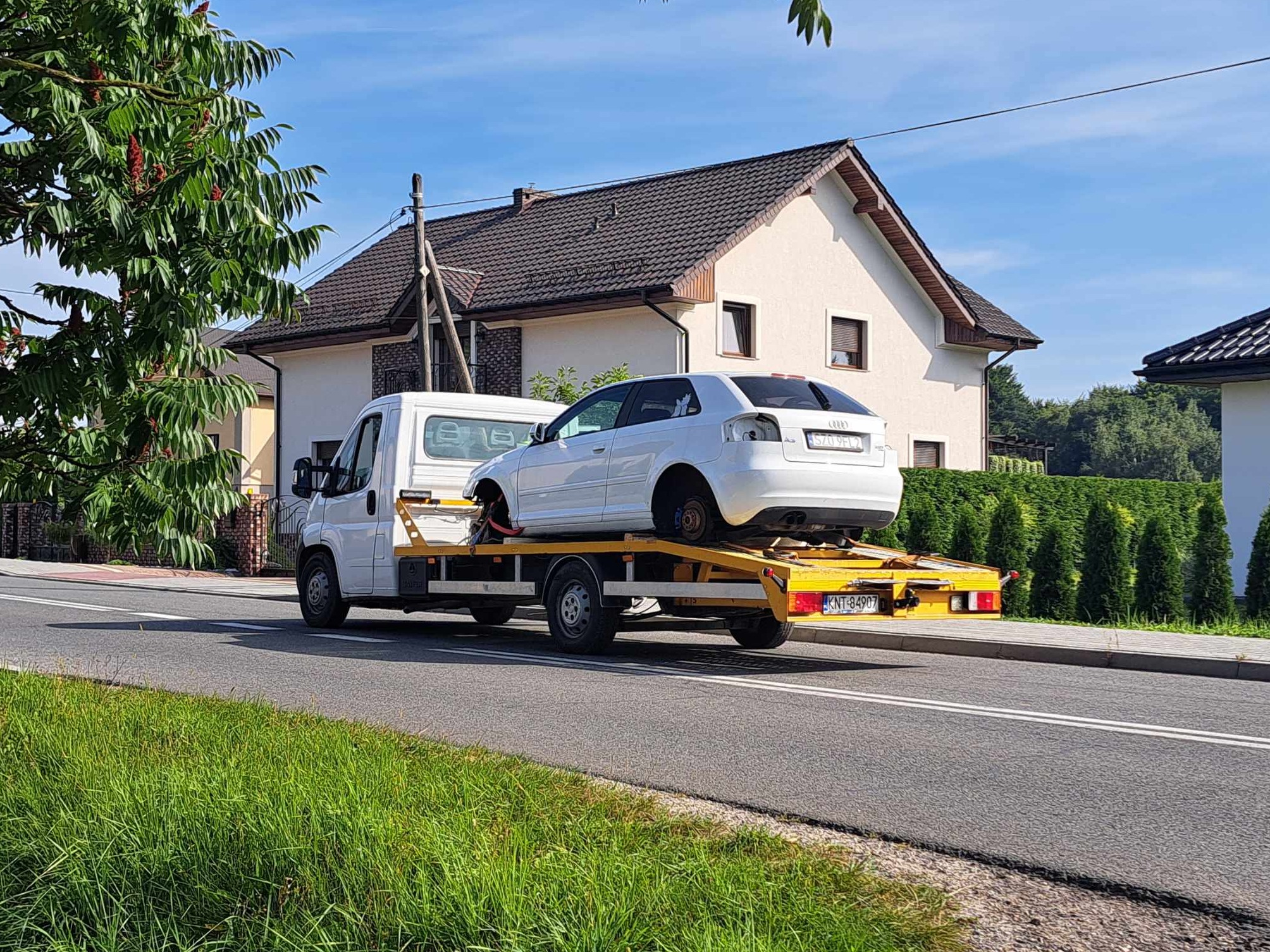 Biały samochód Audi A3 bez kół załadowany na żółtą lawetę na tle jednorodzinnego domu i zieleni, z widoczną tablicą rejestracyjną lawety.