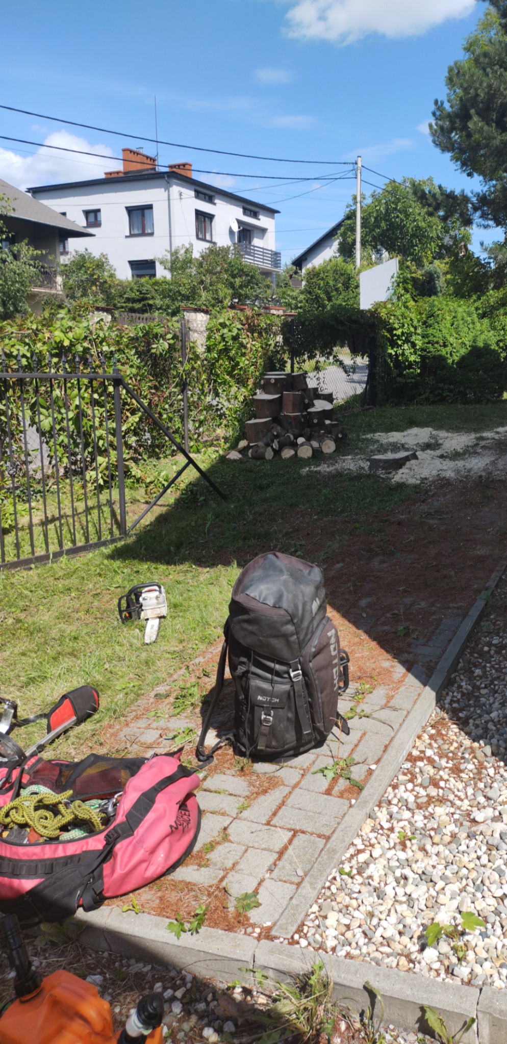 Sprzęt do wycinki drzew rozłożony na trawniku i kostce brukowej: piła łańcuchowa, plecak marki Notch, lina, kanister z paliwem, rękawice. W tle sterta pociętych pni drzew i dom jednorodzinny.