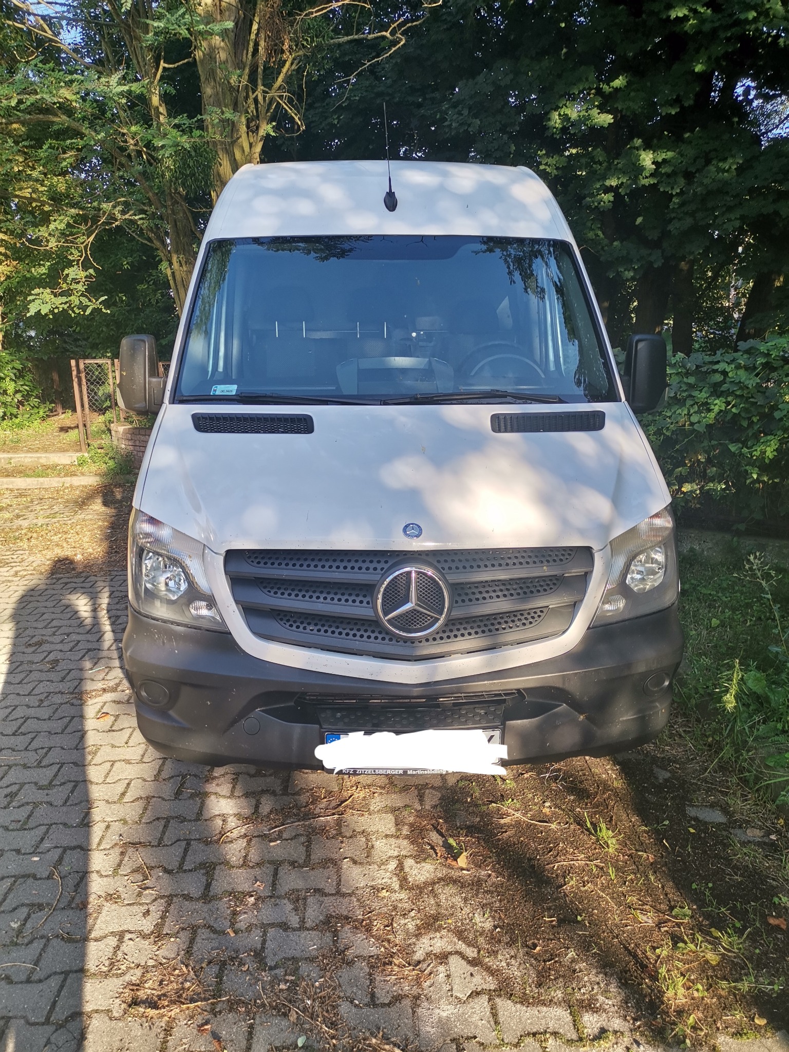 Biały bus marki Mercedes-Benz zaparkowany na brukowanej nawierzchni, z widoczną atrapą chłodnicy z dużym logo i fragmentem tablicy rejestracyjnej.