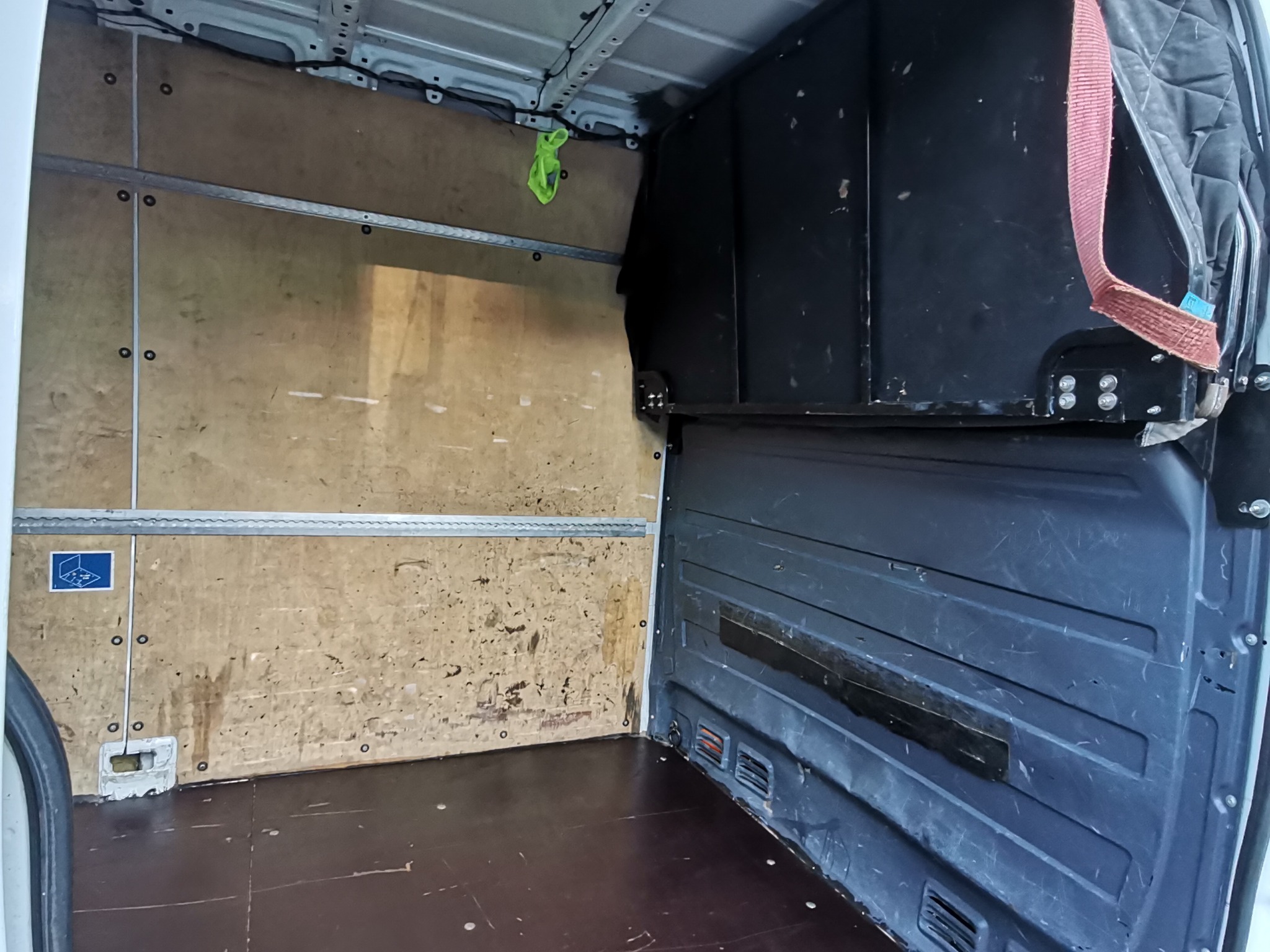 Wnętrze puste furgonetki z drewnianą podłogą i ścianami zabezpieczonymi płytami, widoczne mocowania do zabezpieczenia ładunku, w tle czarna zabudowa ochronna.