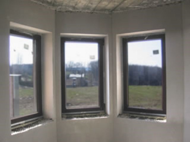 Surowe tynki na ścianach i suficie w pomieszczeniu z trzema oknami, widoczne niedoskonałości i nierówności powierzchni przed wykończeniem.