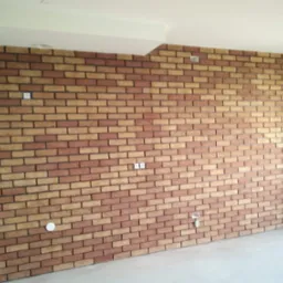 Wnętrze z wykończoną ścianą z cegły klinkierowej w odcieniach brązu i beżu, widoczne gniazdka elektryczne, jasna podłoga i fragment sufitu.