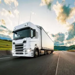 Biała ciężarówka z naczepą w ruchu na autostradzie, z rozmytym tłem gór i zachmurzonego nieba oświetlonego zachodzącym słońcem.