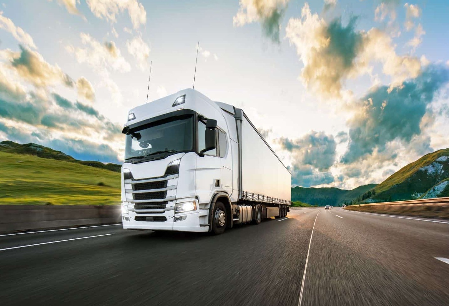 Biała ciężarówka z naczepą w ruchu na autostradzie, z rozmytym tłem gór i zachmurzonego nieba oświetlonego zachodzącym słońcem.