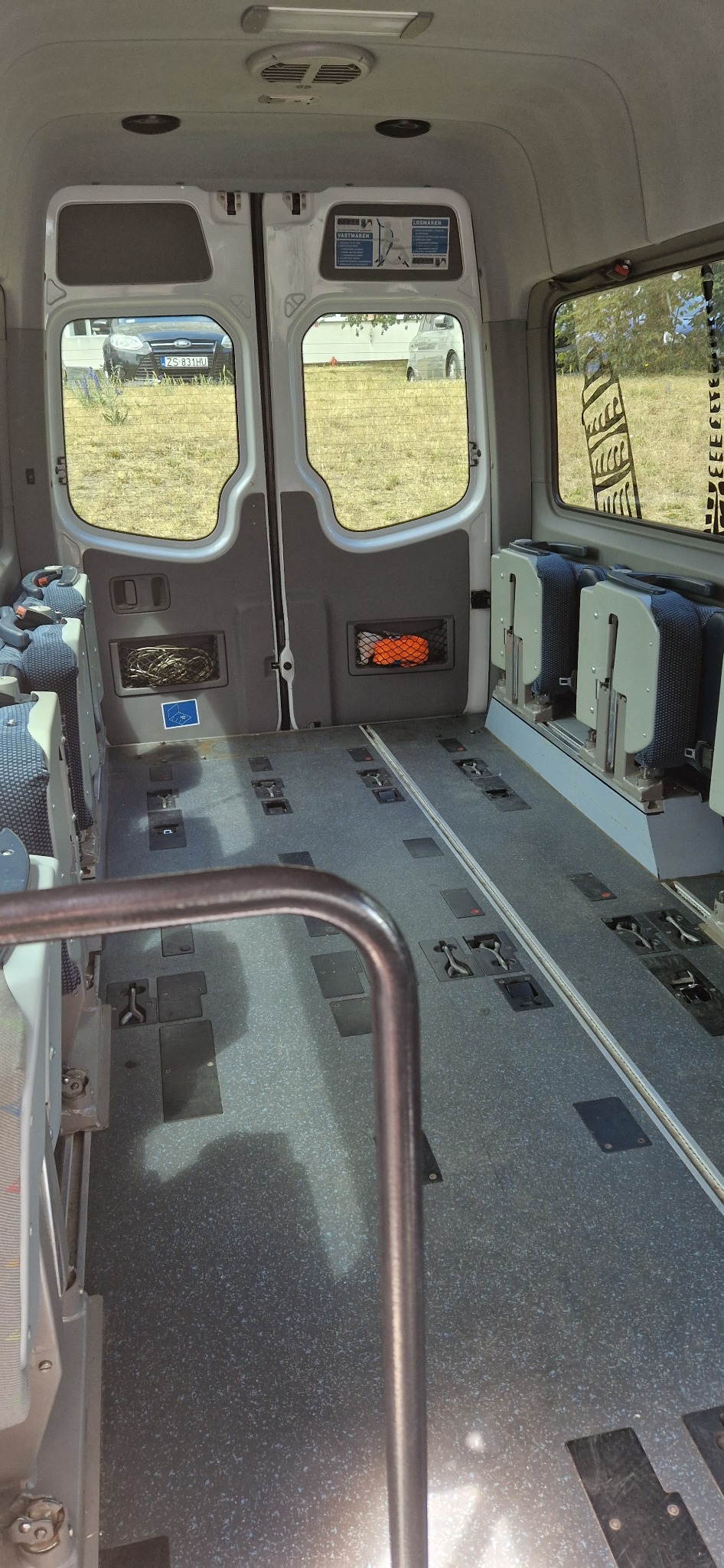 Wnętrze autobusu z demontowalnymi siedzeniami, widoczne szyny montażowe w podłodze, siatki na drobiazgi na tylnych drzwiach.