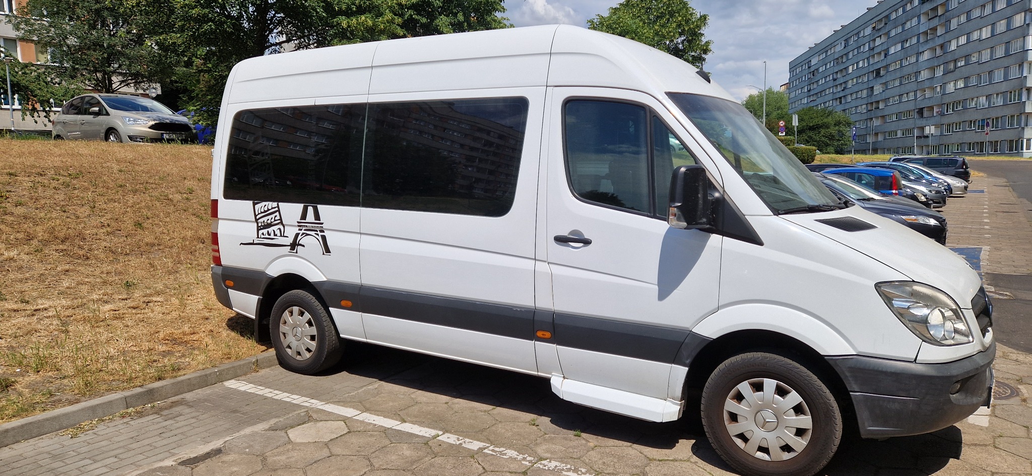 Biały bus zaparkowany na parkingu, z grafiką przedstawiającą Koloseum i Wieżę Eiffla na boku.