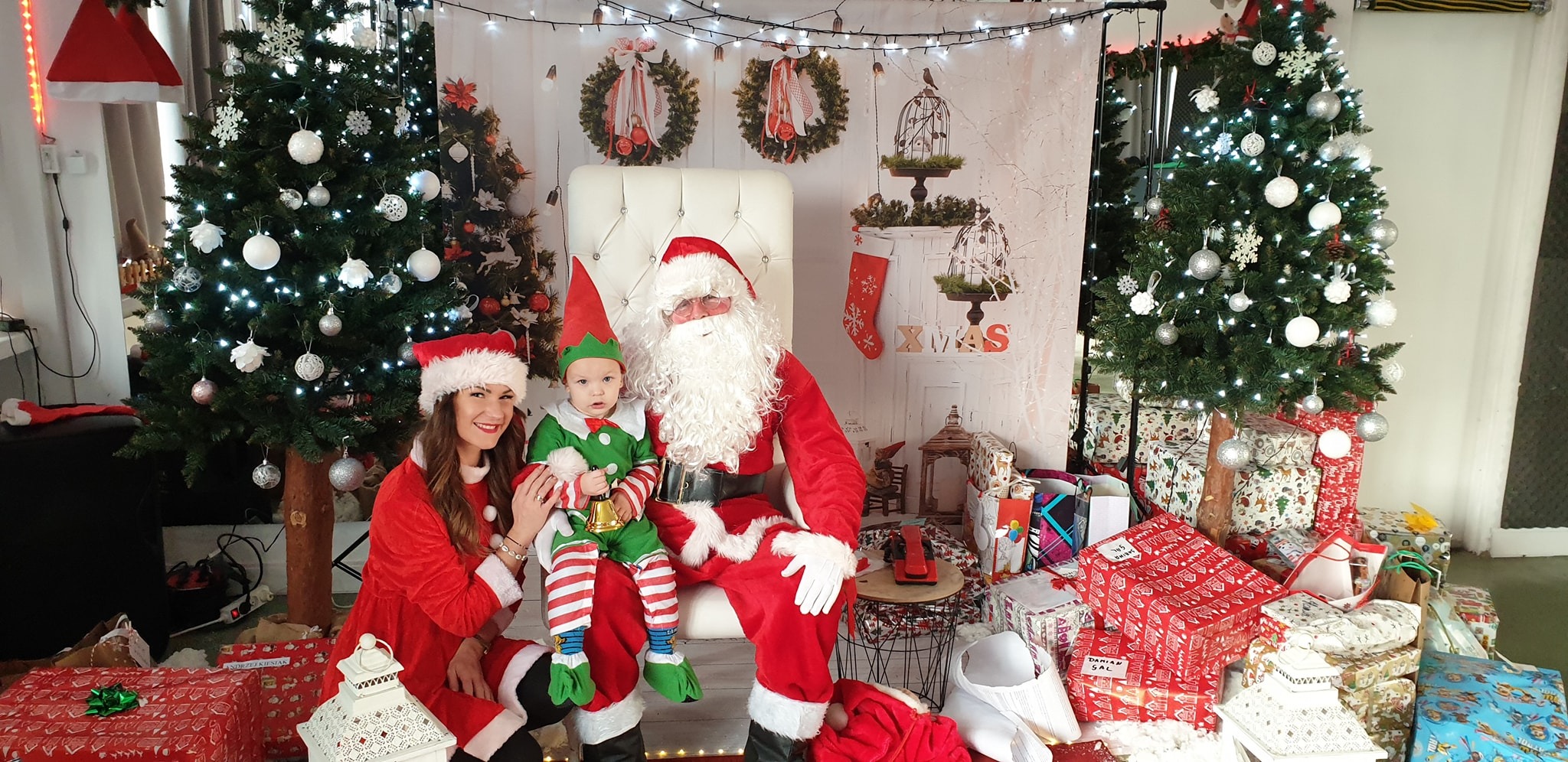 Sesja zdjęciowa z Mikołajem i elfem na tle świątecznej scenografii z choinkami i prezentami.