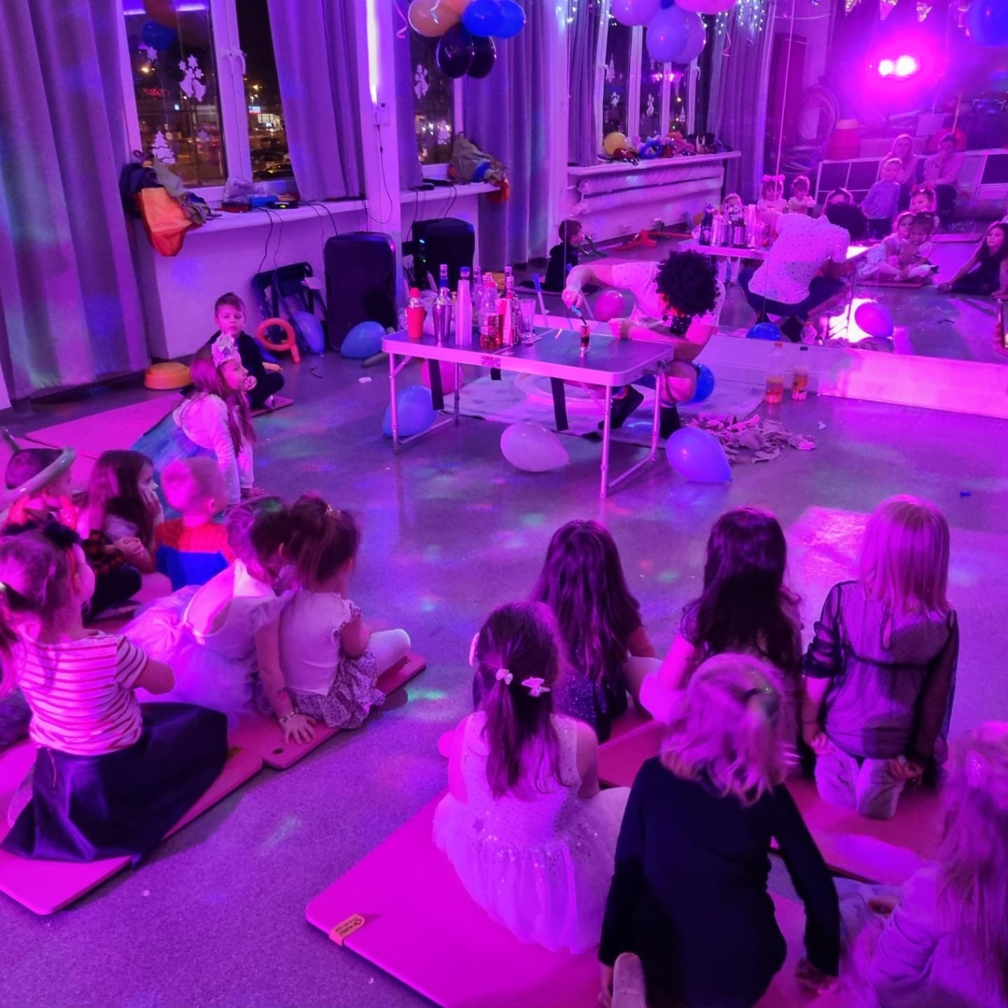 Animator w afro peruce przygotowuje kolorowe napoje dla grupy dzieci siedzących na różowych matach w sali z balonami, oświetlonej fioletowym światłem.