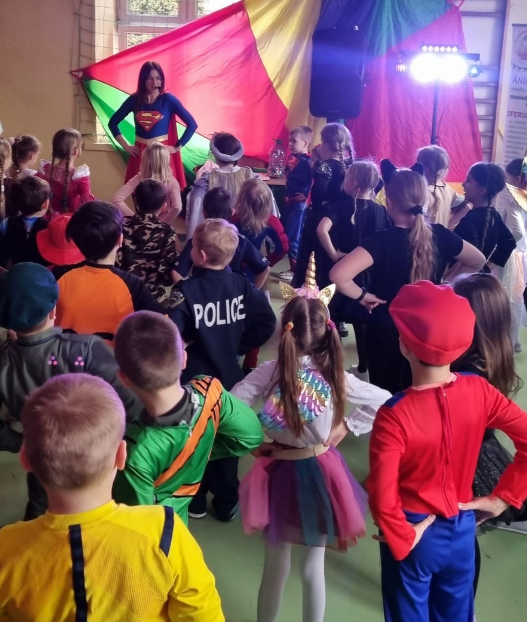 Kobieta w kostiumie superbohatera prowadzi grupę dzieci przebranych w różne stroje, w tle kolorowa dekoracja i oświetlenie sceniczne.