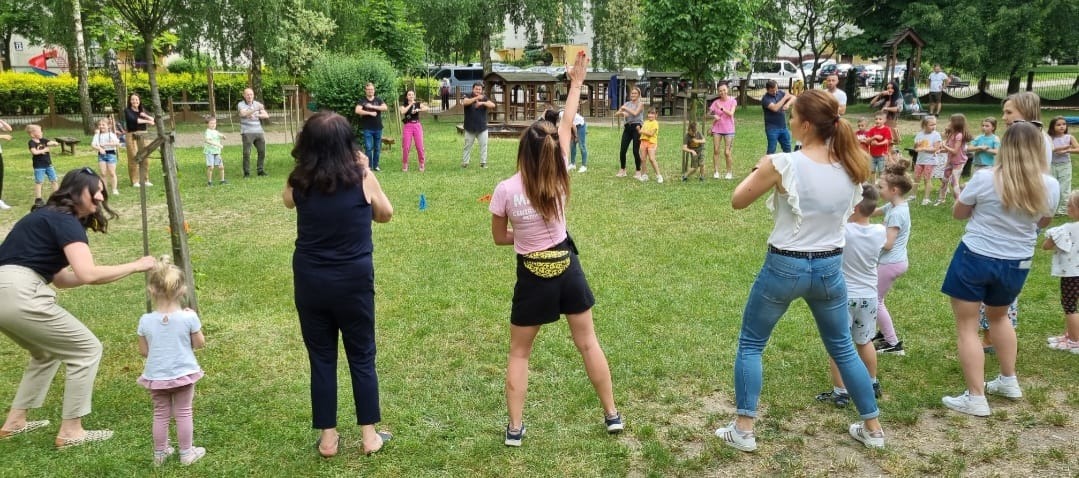 Grupa dzieci i dorosłych na trawniku wykonuje ćwiczenia pod okiem instruktorów, w tle widoczne drzewa i plac zabaw.