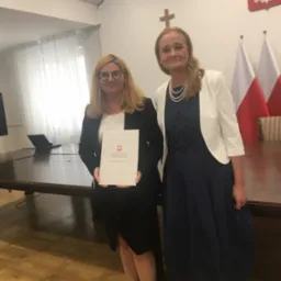 Dwie kobiety w oficjalnych strojach pozują w pomieszczeniu z flagami Polski i godłem państwowym. Jedna trzyma dokument Ministerstwa Sprawiedliwości.