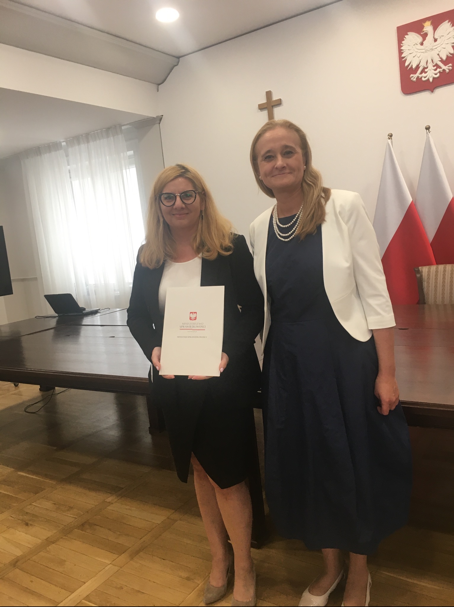 Dwie kobiety w oficjalnych strojach pozują w pomieszczeniu z flagami Polski i godłem państwowym. Jedna trzyma dokument Ministerstwa Sprawiedliwości.