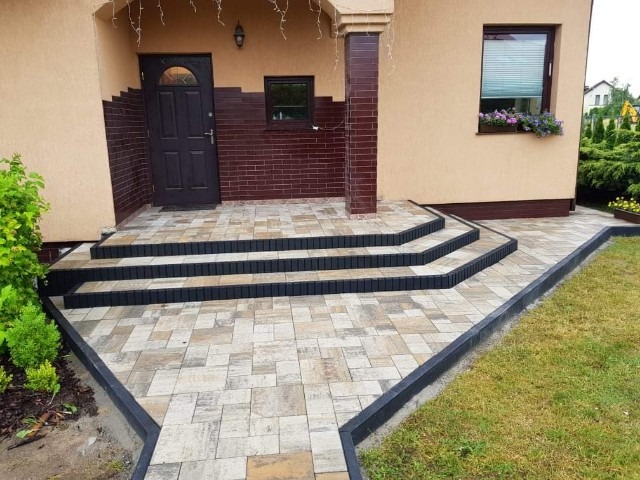 Ułożona kostka brukowa przed domem z trzema schodami, obrzeża z ciemnej kostki, trawnik po prawej stronie, kwiaty w oknie na piętrze.