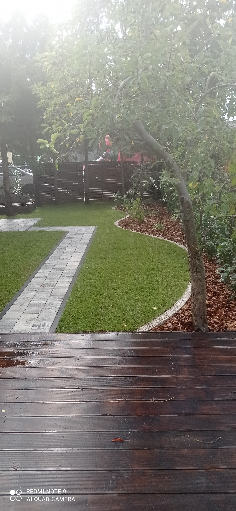 Ogród po deszczu: świeżo ułożony trawnik z kostkową ścieżką, rabaty wyściółkowane korą, widok z mokrego, drewnianego tarasu.