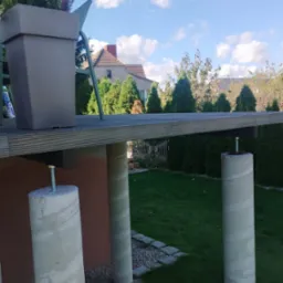 Konstrukcja z betonowymi słupami i drewnianym blatem, widoczna z boku, z donicą na górze, na tle zielonego ogrodu i błękitnego nieba z chmurami.