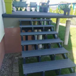 Schody z desek kompozytowych prowadzące na podwyższony taras z meblami ogrodowymi i roślinami w donicach, widok z boku na tle trawnika i drzew